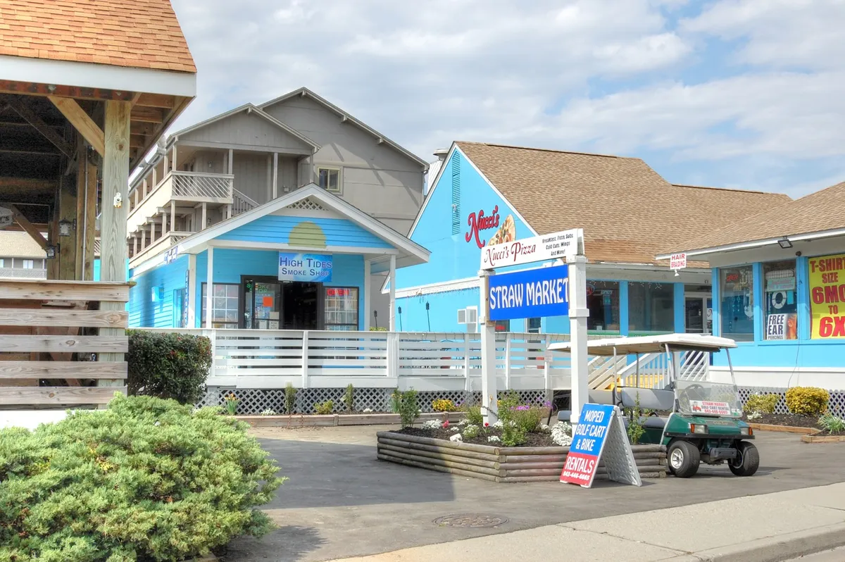 A cheerful street scene with brightly painted buildings, visible signage for the Straw Market and a pizza shop, and a golf cart in the foreground. The setting feels welcoming and hints at the vibrant local dining and shopping scene accessible from the condo.
