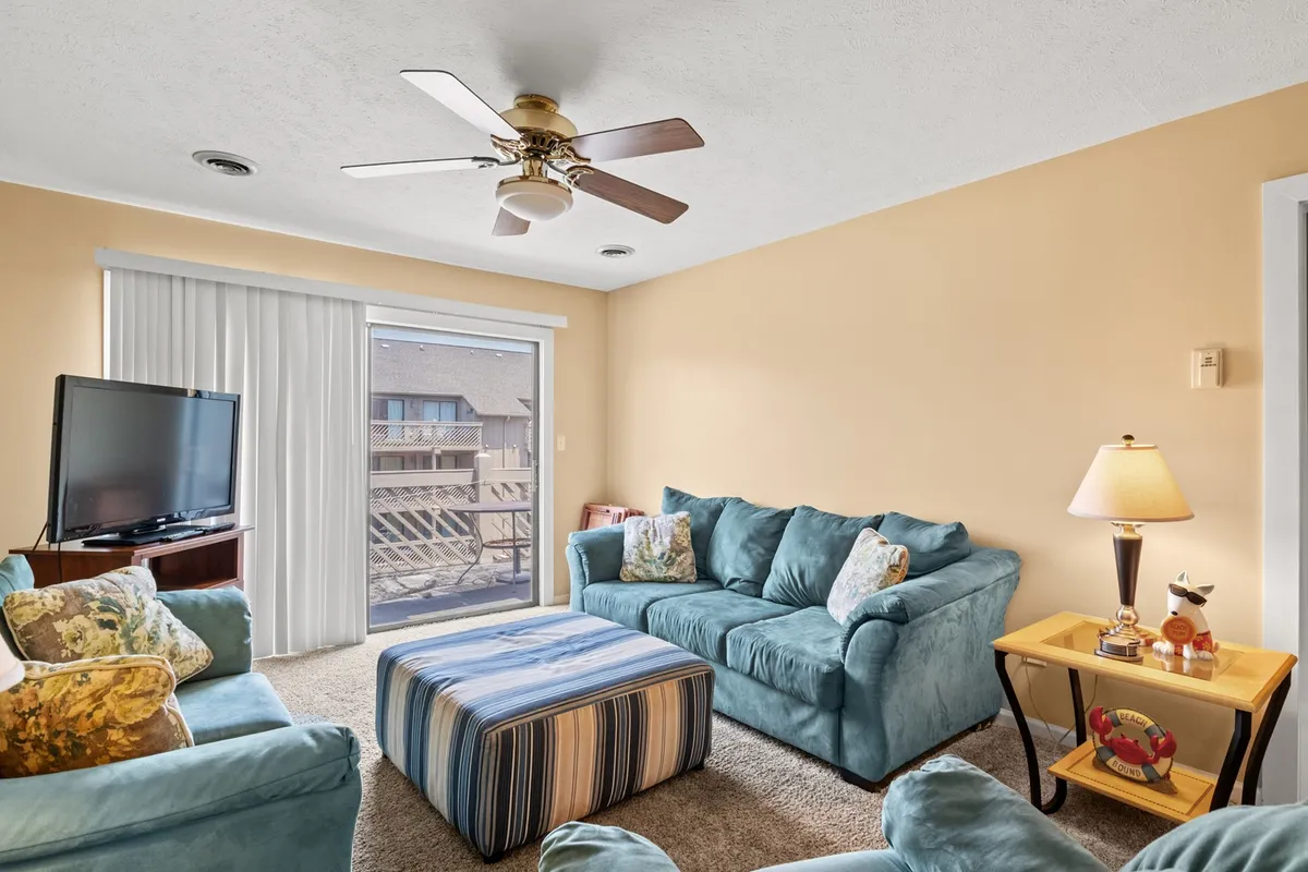 This image showcases a comfortable, open living room with a coordinated sofa set, a striped ottoman, and a television, all arranged around a bright sliding glass door that opens to a private balcony. Natural light pours in, enhancing the coastal décor and inviting atmosphere.