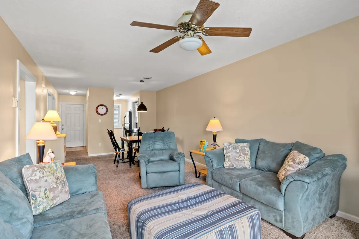 The photo reveals a broader view of the living space, featuring sofas, a coffee table, lamps, and a visible dining area in the background. The layout encourages flow between relaxing and dining, underscoring the open-concept design.