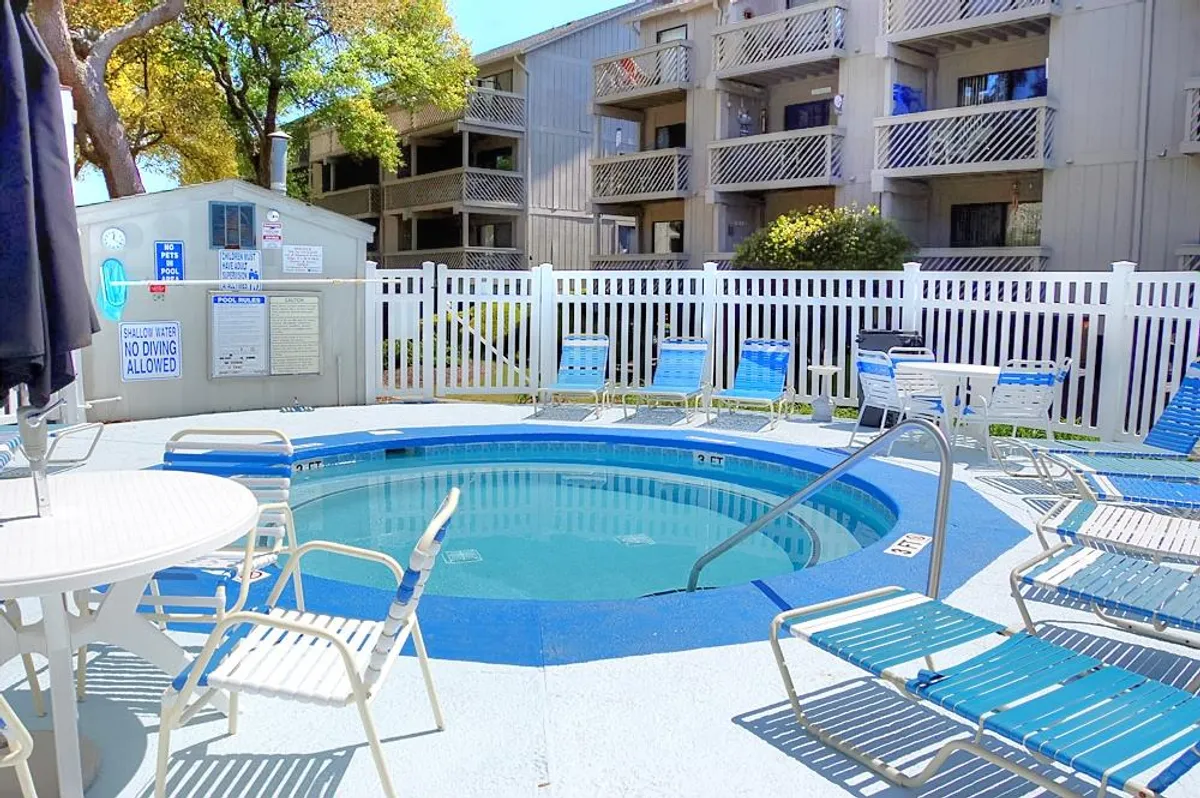 A sunlit pool area featuring a hot tub, surrounded by greenery and comfortable lounge chairs within a fenced space. The atmosphere is relaxed and inviting, with plenty of room for groups to gather, unwind, and enjoy the outdoors together.
