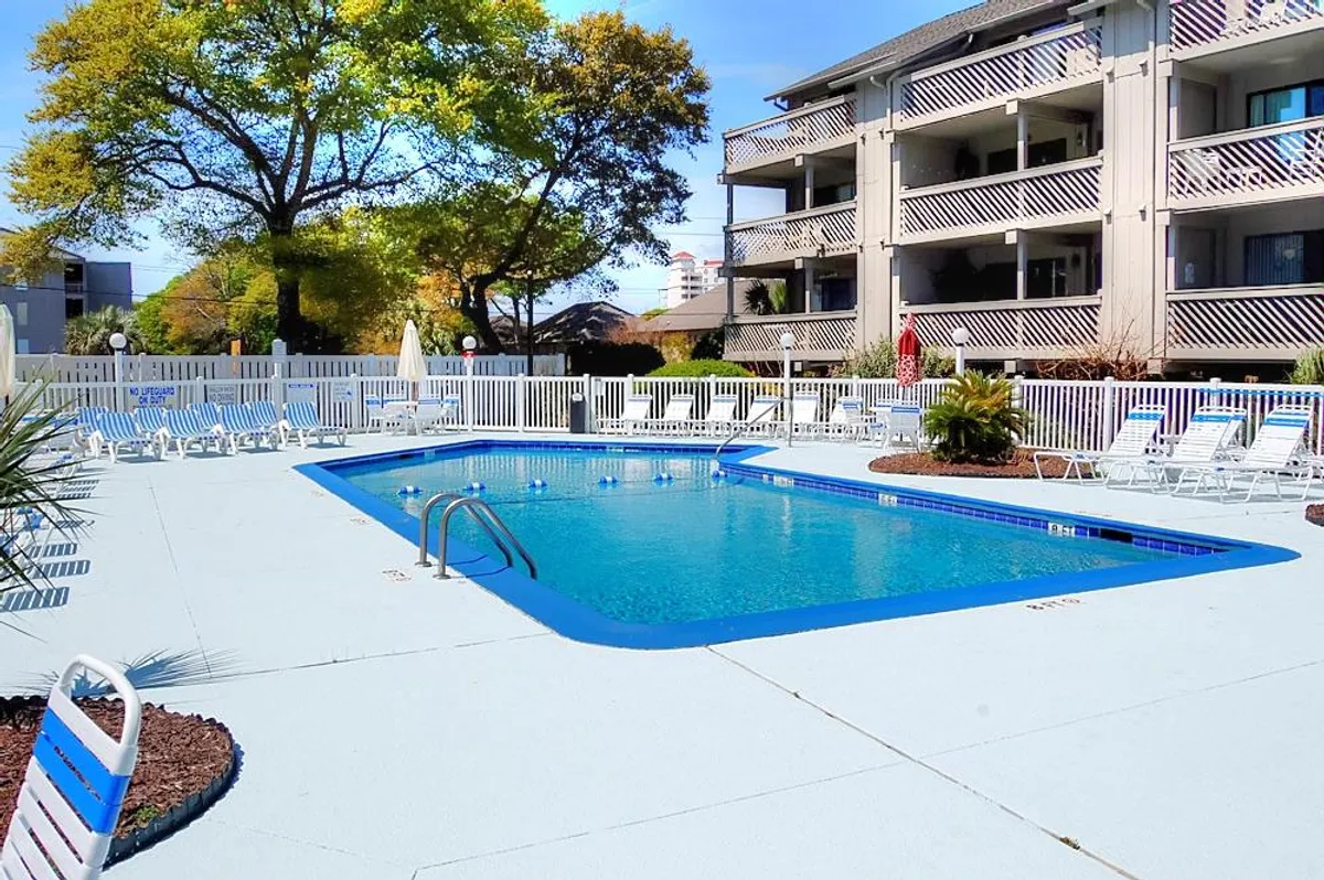 The image showcases the inviting outdoor pool area at Shipwatch Pointe II, bordered by lounge chairs and lush trees, with the building's balconies overlooking the vibrant scene. Guests can be seen swimming, sunbathing, and socializing, set against a bright, sunny Myrtle Beach backdrop.