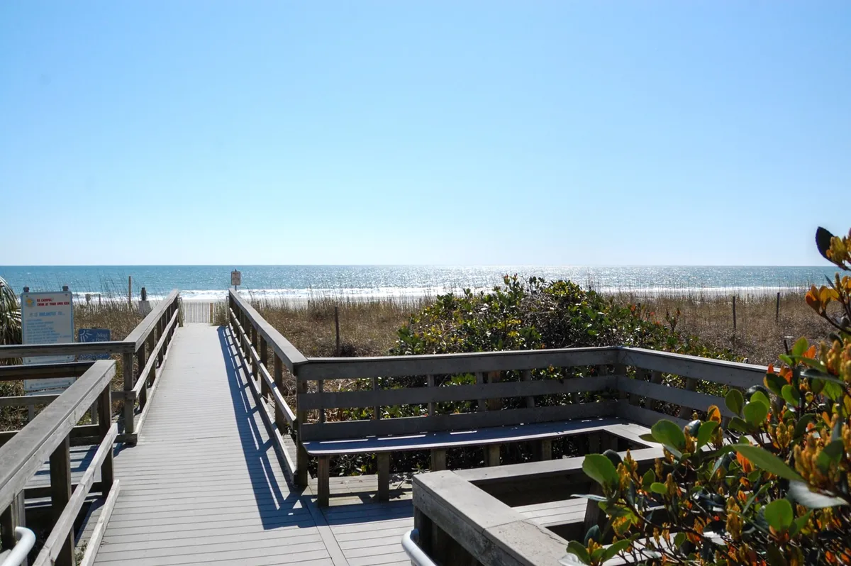 A picturesque wooden walkway leads through beach grass and dune vegetation, opening up to an expansive view of the ocean just across the street. The tranquil setting invites activities like walking to the beach, sunbathing, swimming, and building sandcastles.