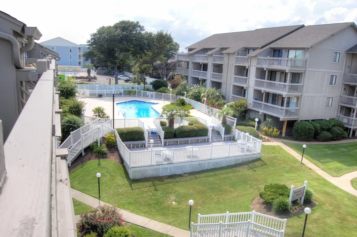 A spacious outdoor swimming pool set amidst landscaped gardens, with sun loungers and the balconies of the resort buildings visible in the background. The calm water, green surroundings, and inviting poolside environment create an atmosphere of relaxation and recreation.