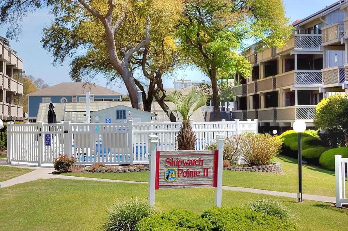 The Shipwatch Pointe II entrance sign stands near a sparkling swimming pool surrounded by lush greenery and neighboring buildings, evoking a welcoming resort atmosphere. The pool is inviting, with loungers perfect for family relaxation or social gatherings.