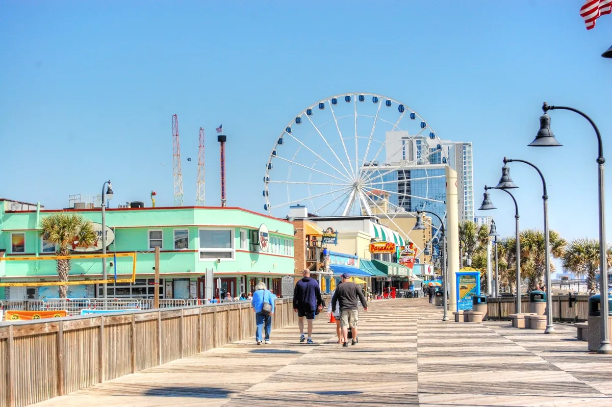 A lively exterior scene of a bustling boardwalk with a ferris wheel, colorful buildings, and people strolling, shopping, and enjoying amusement rides. The atmosphere is joyful and energetic, reflecting popular local attractions for guests of all ages.