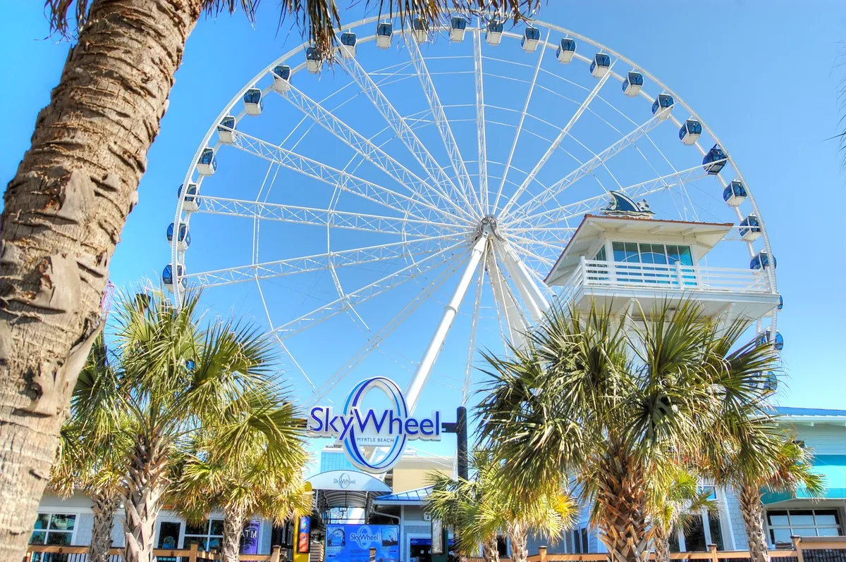 The iconic SkyWheel Ferris wheel rises above palm trees, with the SkyWheel entrance sign in view. The lively scene hints at festive outings, panoramic views, and family-friendly adventure right on the Myrtle Beach boardwalk.