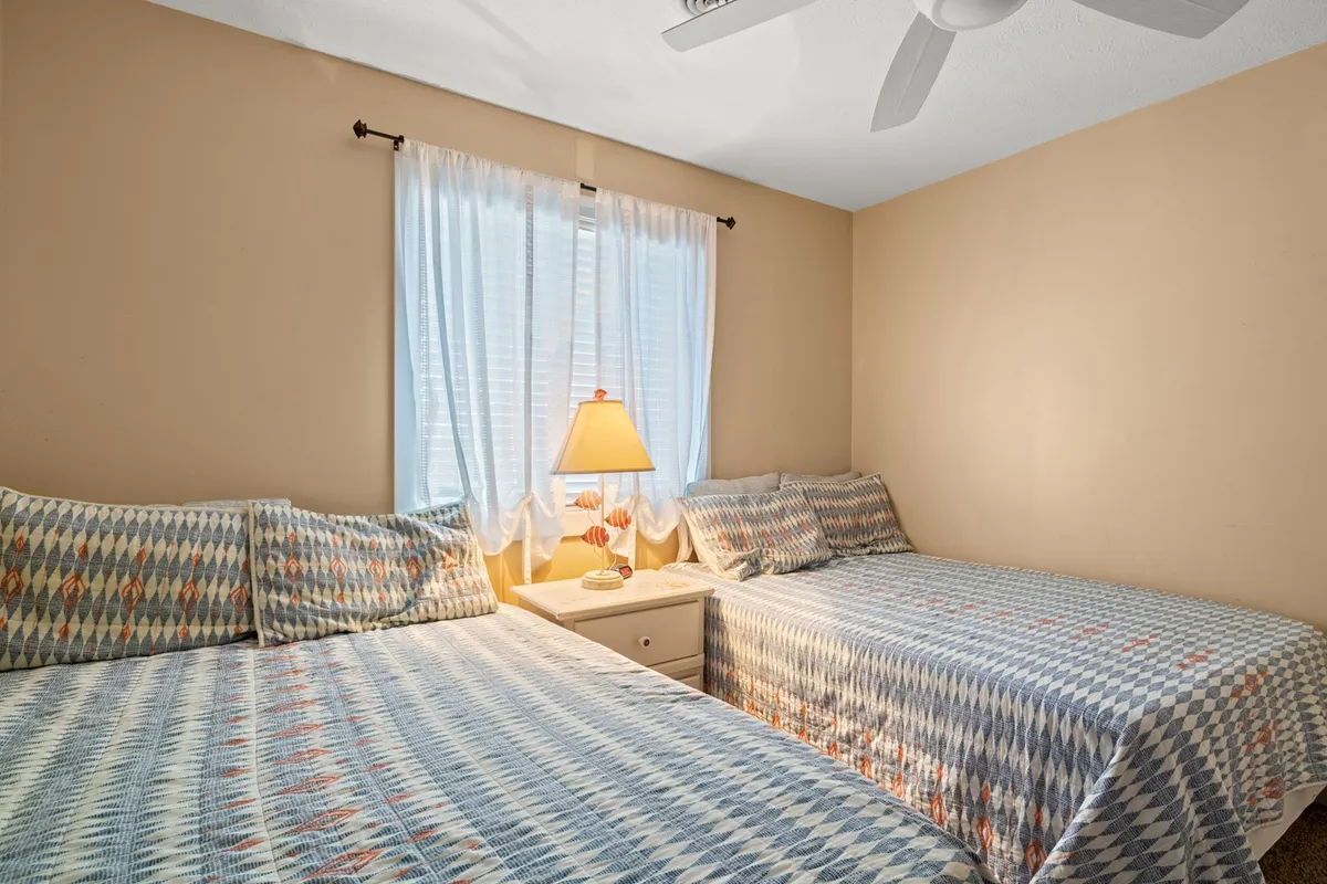 A bright second bedroom with two neatly made beds, a window with light curtains letting in the morning sun, a central lamp providing warm illumination, and minimalistic decor that keeps the space open and welcoming.